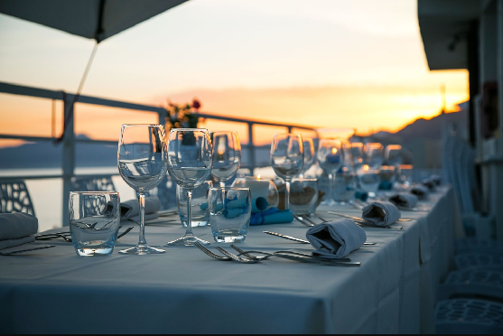 A long outdoor restaurant terrace table covered in a white tablecloth, formally set with water glasses, wine glasses, and folded napkins, overlooking a calm sea and distant mountains during a bright orange sunset.